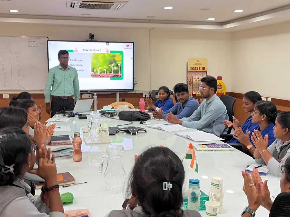 A facilitator presenting to a small group seated around a table, a slide on the screen behind him